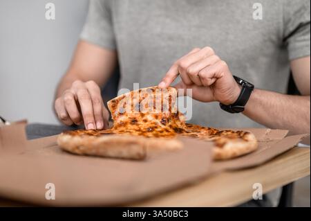 Arab man taking a slice of pizza at home in a kitchen setting. A casual indoor food scene ideal for lifestyle, comfort food, and nutrition marketing. Great for promotions around meals, delivery, and home dining. Stock Photo