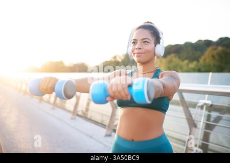 A dark-eyed woman wearing earphones lifts dumbbells in an outdoor workout. She focuses on building muscle and vitality through strength training, perfect for fitness and wellness campaigns. Stock Photo