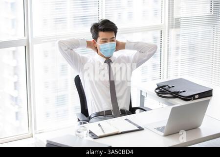 Smiling Chinese businessman wearing a face mask sits at his desk with a laptop. A professional office scene that reflects resilience and safety during the Covid era. Ideal for business, health, and workplace communications focusing on well-being and online work. Stock Photo
