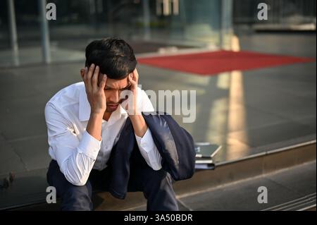 Stressed millennial Asian businessman sits on the stairs outside a company building, expressing frustration after a career setback. The image conveys workplace stress and challenges in modern business life, suitable for articles on unemployment, job loss, and resilience. Stock Photo