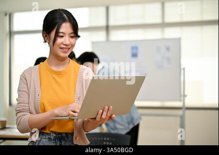 A stylish young Asian female graphic designer stands in a modern co-working office with a laptop in hand. This professional portrait highlights creativity and tech-savvy website design in a collaborative workspace. Ideal for topics on UI and UX design, web development, startups and remote work. Stock Photo