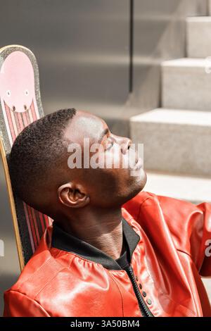 Side view of a stylish young Black man in his 20s, wearing a leather jacket and leaning on a skateboard. He relaxes on a sunlit staircase with his hands behind his head and eyes closed, exuding calm confidence. This urban street style image works well for lifestyle, youth culture and fashion campaigns. Stock Photo