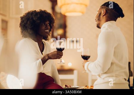 A smiling Black couple shares a romantic dinner at home, chatting and sipping wine. Candlelight and an elegant table setting create a warm, intimate atmosphere suitable for romance, couples photography and home lifestyle themes. Stock Photo