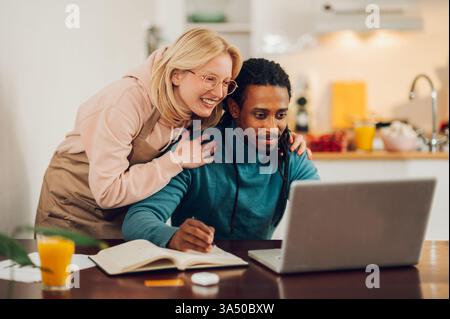 A happy multicultural couple enjoy a moment at home, with the husband following an online lecture on a laptop. His partner offers warm support, capturing a caring, connected dynamic. This scene highlights digital learning, home life and modern technology in everyday life. Stock Photo