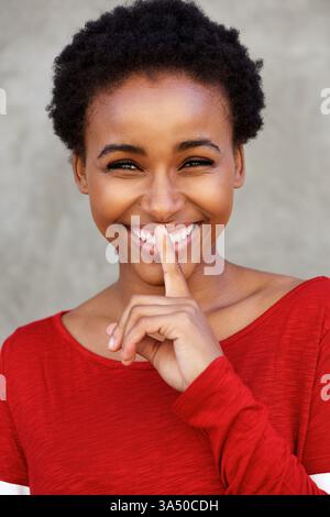 Portrait of a cute young woman smiling with her finger over her mouth in a shushing gesture. The close-up captures a playful, confident mood, suitable for lifestyle and fashion content. Stock Photo