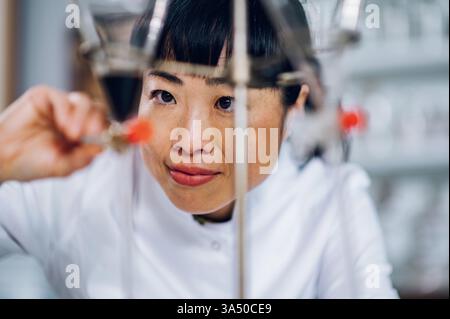 Portrait of a Japanese pharmacist in a vintage apothecary, handling a glass lab vessel for medicine preparation. The focused professional wears a lab coat in a studio setting, showcasing pharmaceutical work and healthcare expertise. Ideal for healthcare, pharmacy and medical industry themes. Stock Photo