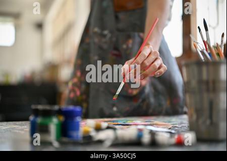 Close-up of a gay Asian artist in an apron dipping a paintbrush into watercolor, creating artwork in the studio. This vibrant, creative scene is ideal for art, design, and LGBTQ inclusive campaigns. Stock Photo