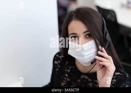 Hispanic female call center operator wearing headset and mask at her desk in an office setting. This image suits customer service, frontline roles, and workplace safety during the pandemic. Stock Photo