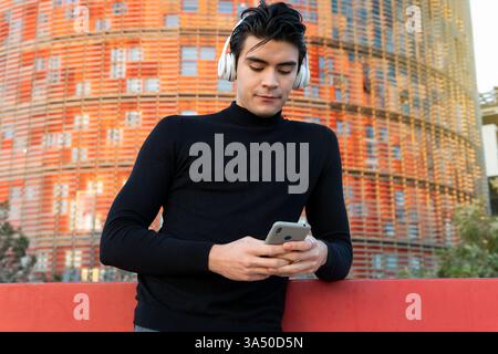 Hispanic man wearing headphones and using a smartphone on a city street. A modern urban lifestyle image suitable for music, mobile apps, and tech connectivity campaigns. Stock Photo