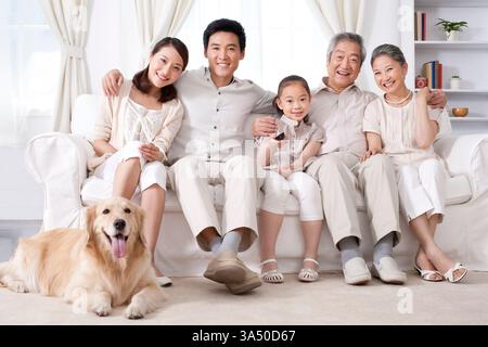 A cheerful Chinese family relaxes at home in a modern living room, watching television together with a dog nearby. This scene highlights family life, togetherness and domestic bliss. Stock Photo