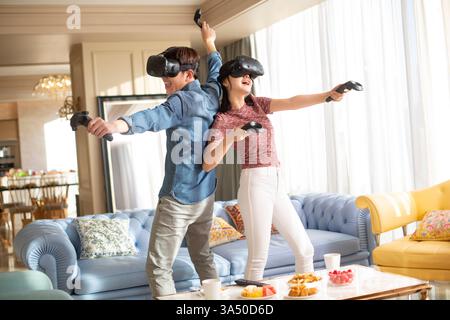 Cheerful Chinese couple enjoy a virtual reality game at home, sharing a playful moment in a cozy living room. This lifestyle image captures togetherness, gaming and contemporary home entertainment. Stock Photo
