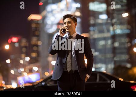 Smiling Chinese businessman on a night city street, talking on his phone as he walks away from a parked car. An urban moment ideal for business, city life, and modern work scenarios. Stock Photo