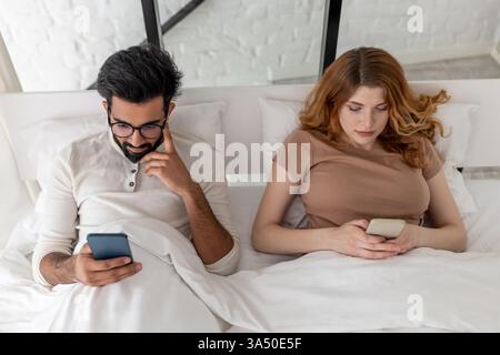 Young mixed-race couple lying on bed using a smartphone with serious expressions. The couple stays focused as they text on a smartphone in a bedroom setting. A contemporary lifestyle image that captures digital communication and relationship dynamics. Stock Photo