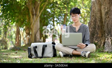 Handsome young Asian male college student wearing glasses studies on campus, sipping coffee while using a laptop. A relaxed, focused moment suitable for education and student life campaigns. Great for campus tech, cafe culture, and academic branding. Stock Photo