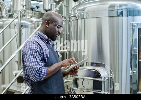 Serious African American engineer using a digital tablet in a brewery workshop. He stands among stainless steel equipment and factory machinery, focused on the task at hand. Ideal for content about industrial technology, beer production, and manufacturing careers. Stock Photo