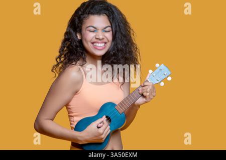 Smiling African American woman playing a ukulele while standing against a wall. Great for music, hobby and lifestyle visuals that convey joy and creativity. Ideal for campaigns celebrating relaxed leisure and musical moments. Stock Photo