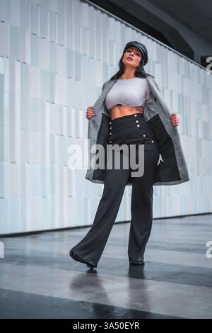 Stylish Latin woman posing in the city wearing a leather hat and a gray trench coat. Urban fashion portrait with autumn styling and a confident expression. Ideal for street style features, fashion editorials, and city lifestyle content. Stock Photo