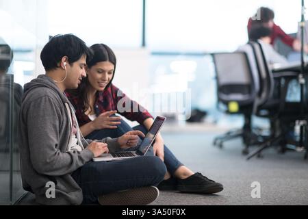 Smiling Mixed race man with earphones using laptop with caucasian female coworker sitting on floor in office Stock Photo