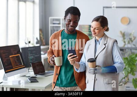 Smiling businesswoman with a bionic arm shows a photo on a smartphone to a colleague during a break. The image emphasizes disability inclusion and assistive technology in the workplace. Ideal for stories about teamwork, innovation, and adaptive equipment in corporate settings. Stock Photo