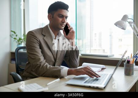 Portrait of a focused Indian entrepreneur in a busy office, talking on a phone while checking emails and documents on a laptop. This image suits business, technology, and startup themes. Ideal for reports, presentations, and corporate communications. Stock Photo