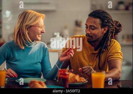 Happy interracial couple enjoying a breakfast at home, sharing a croissant. Cozy, sunlit dining area mood with warmth and affection. Great for lifestyle, relationships, and multicultural branding. Stock Photo