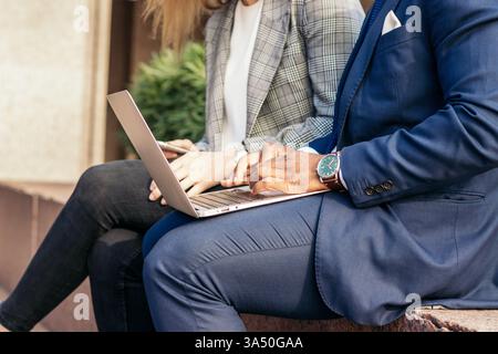 Business colleagues in suits working on a laptop outdoors. Professional team scene showing collaboration in an outdoor setting. Ideal for corporate, technology, and productivity content. Stock Photo