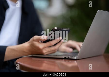 Cropped image of a young Asian man working remotely in a coffee shop, using a smartphone and a laptop. This scene is ideal for remote work, digital nomad and urban lifestyle themes. Stock Photo