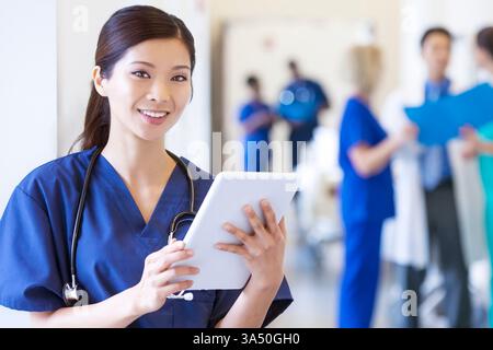 Smiling Chinese nurse in scrubs using a wireless tablet at a modern medical center. This healthcare portrait highlights digital health and patient care, ideal for hospital and medical marketing. Stock Photo