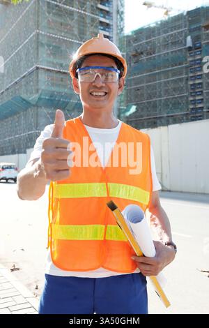 Happy head engineer stands at an outdoor construction site, showing thumbs up beside a level and rolled blueprint. Dressed in protective workwear, he embodies professionalism in the construction and engineering field. Great for construction, architecture, and project planning visuals. Stock Photo