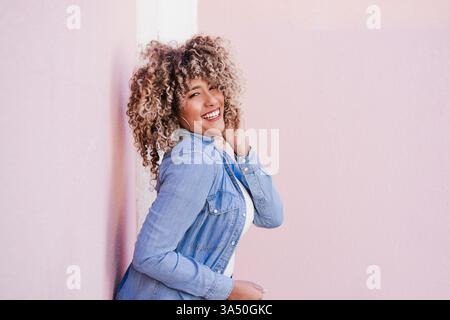 Cheerful Hispanic woman leaning against a pink wall, smiling and touching her curly hair. A bright lifestyle portrait suitable for fashion, beauty, and everyday positivity. Stock Photo