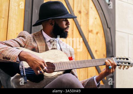 Stylish Black man in a hat and classic outfit plays guitar outdoors. The image captures a joyful musician in a natural setting, perfect for music and lifestyle branding. Suitable for campaigns around outdoor performances and cultural music. Stock Photo