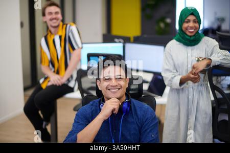 Diverse startup team in a modern open plan office, featuring an Arabian woman wearing a hijab during a brainstorming session. The group collaborates around a laptop and desktop, showcasing teamwork and contemporary workplace culture. Great for entrepreneurship, diversity in business, and modern offices. Stock Photo