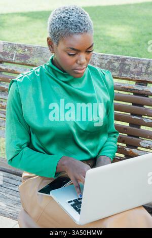 Black woman works on a laptop while seated on a wooden bench in the park. This remote work setting highlights focus, outdoor productivity, and contemporary work-life balance. Stock Photo