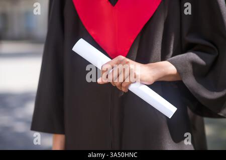 African American female in graduation gown holding diploma standing outdoors during daytime. A proud graduate moment suitable for education, achievement, and student success campaigns. Perfect for post-graduation opportunities and university promotion. Stock Photo