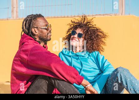 A smiling multiracial couple sits on the ground in a sunny urban setting. They wear hoodies and sunglasses and share a cheerful moment looking at each other. Ideal for lifestyle and relationship features highlighting diverse urban life. Stock Photo