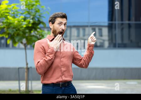A surprised man in a shirt points upwards with an open mouth and a hand to his mouth, standing outdoors with a modern building behind. Stock Photo