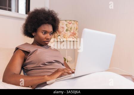Black woman sitting on a bed at home using a laptop for remote work. The relaxed, casual setting suits telecommuting, online learning, and home office themes. Stock Photo