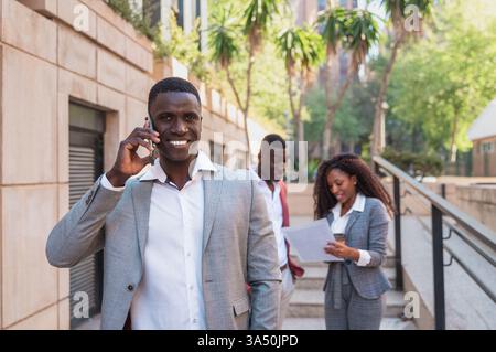 Smiling Black businessman having phone conversation standing near colleagues outside building in city during daytime Stock Photo
