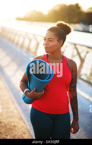 Dark-skinned young woman outdoors in the morning, walking with a sport mat for a workout. Her energetic stride and bright mood suggest motivation and fitness. Ideal for articles on active lifestyle, morning exercise, and wellness. Stock Photo