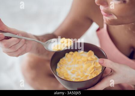 Mixed-race woman with vitiligo eating cereals while relaxing on a bed at home. Great for inclusive lifestyle imagery, wellbeing, and everyday breakfast scenes. Stock Photo
