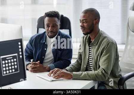Two young programmers from diverse backgrounds work together, entering data at a desk in a modern office. They wear casual clothes in an open space bureau, collaborating during a meeting. Great for articles on teamwork, diversity in tech and workplace collaboration. Stock Photo