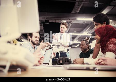 Diverse group of business professionals seated at a table with gadgets and documents, discussing drone technology and VR glasses for business innovation. Perfect for corporate meetings, technology, and startup branding. Stock Photo