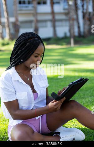 Black woman with braided hair using a tablet while sitting on grass in a park. Casual outdoor lifestyle image suitable for education, remote learning, and digital study. Ideal for content about students, online classes, and university life. Stock Photo