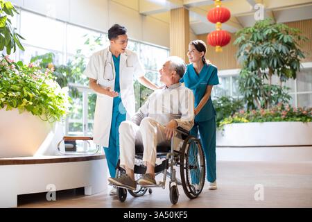 Smiling Chinese doctor talks with a senior patient in a wheelchair, pushed by a nurse in a nursing home. The scene conveys compassionate healthcare, teamwork, and elder care. Suitable for healthcare, aging, and medical professionals imagery featuring Chinese ethnicity. Stock Photo