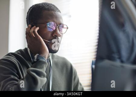 Portrait of a Black man wearing a headset, delivering IT support to a client from a modern office. The scene conveys professional customer service, online help and tech industry communication. Ideal for technology support services, software companies and help desk marketing. Stock Photo