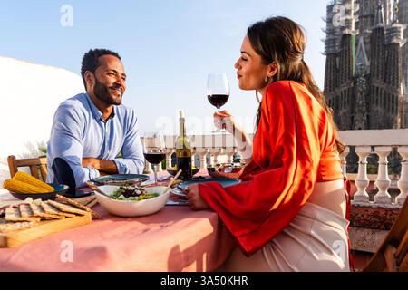 Multiracial couple on a rooftop balcony with views of Sagrada Familia in Barcelona, sharing a romantic aperitif. A sunny travel-inspired scene that conveys love, togetherness, and leisure. Stock Photo