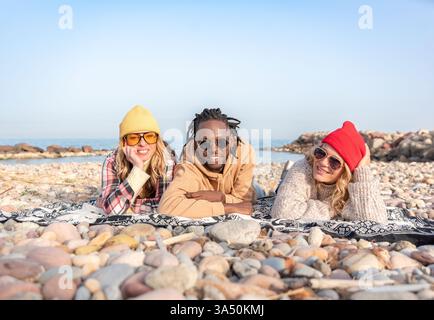Smiling Black man with Caucasian friends lying on a blanket at the beach during summer. A relaxed outdoor lifestyle scene for family, friendship, and vacation mood. Great for summer travel, leisure, and seaside marketing. Stock Photo