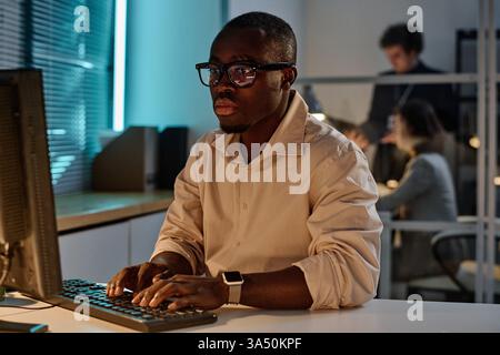 African American programmer concentrating on his online work on a computer in a late evening office. The scene highlights coding, software development, and IT industry themes. Ideal for tech, cybersecurity, and workplace visuals. Stock Photo