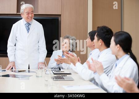 A group of Chinese medical experts clapping in a modern hospital board room, celebrating a successful collaboration. The team includes doctors in lab coats and scrubs, seated around a conference table with a laptop visible. The scene conveys teamwork, leadership, and achievement in healthcare. Stock Photo