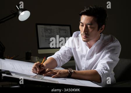 Chinese architect sits at a desk in a contemporary office, studying construction plans. He reviews blueprints and design drawings with pencil and ruler nearby, with a computer screen illuminated in the background. Stock Photo